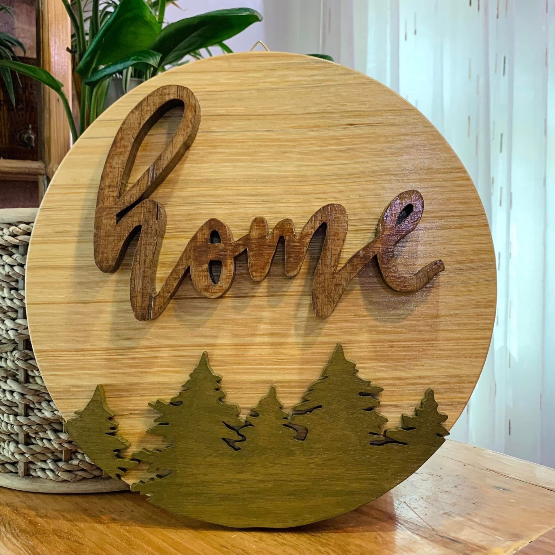 Round light wood sign with brown script 'home' above green pine tree cut-outs